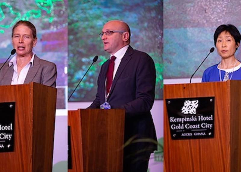 three individuals stand behind a podium, with a large screen displaying information in the background.
