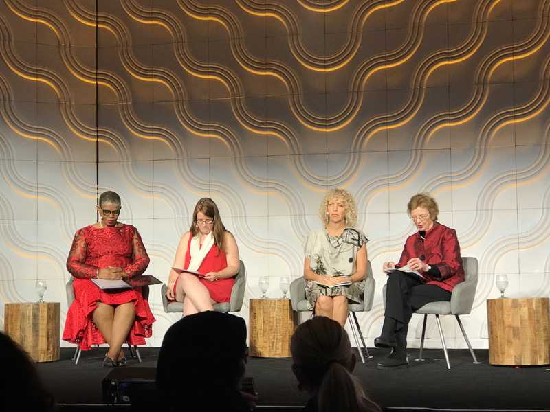 Four women sitting on chairs in front of a stage, discussing tackled women solution.