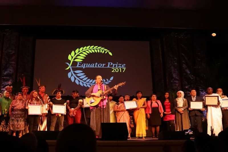 A group of individuals on stage, featuring a woman prominently holding a guitar, engaging the audience with their performance.